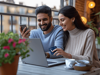 A man and woman sitting together on a balcony, looking at a smartphone and smiling. The woman is holding a Travelex Money Card, suggesting they might be shopping online. A laptop is open in front of them, and there is a coffee cup on the table. A flowering plant adds a touch of nature to the cozy setting.